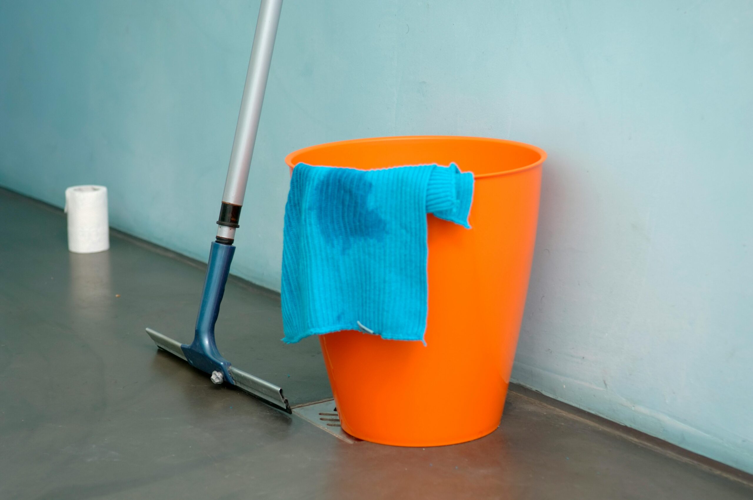 Close-up of cleaning supplies including orange bucket, mop, and blue rag indoors.