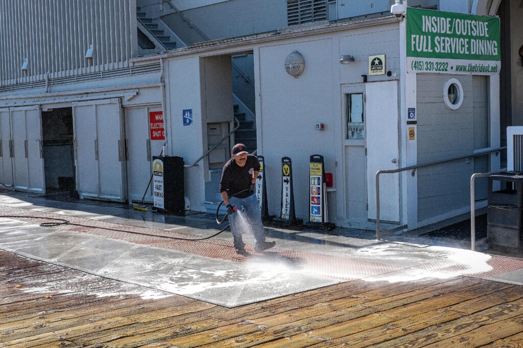 A worker uses a pressure washer to clean a sidewalk outside a dining facility.