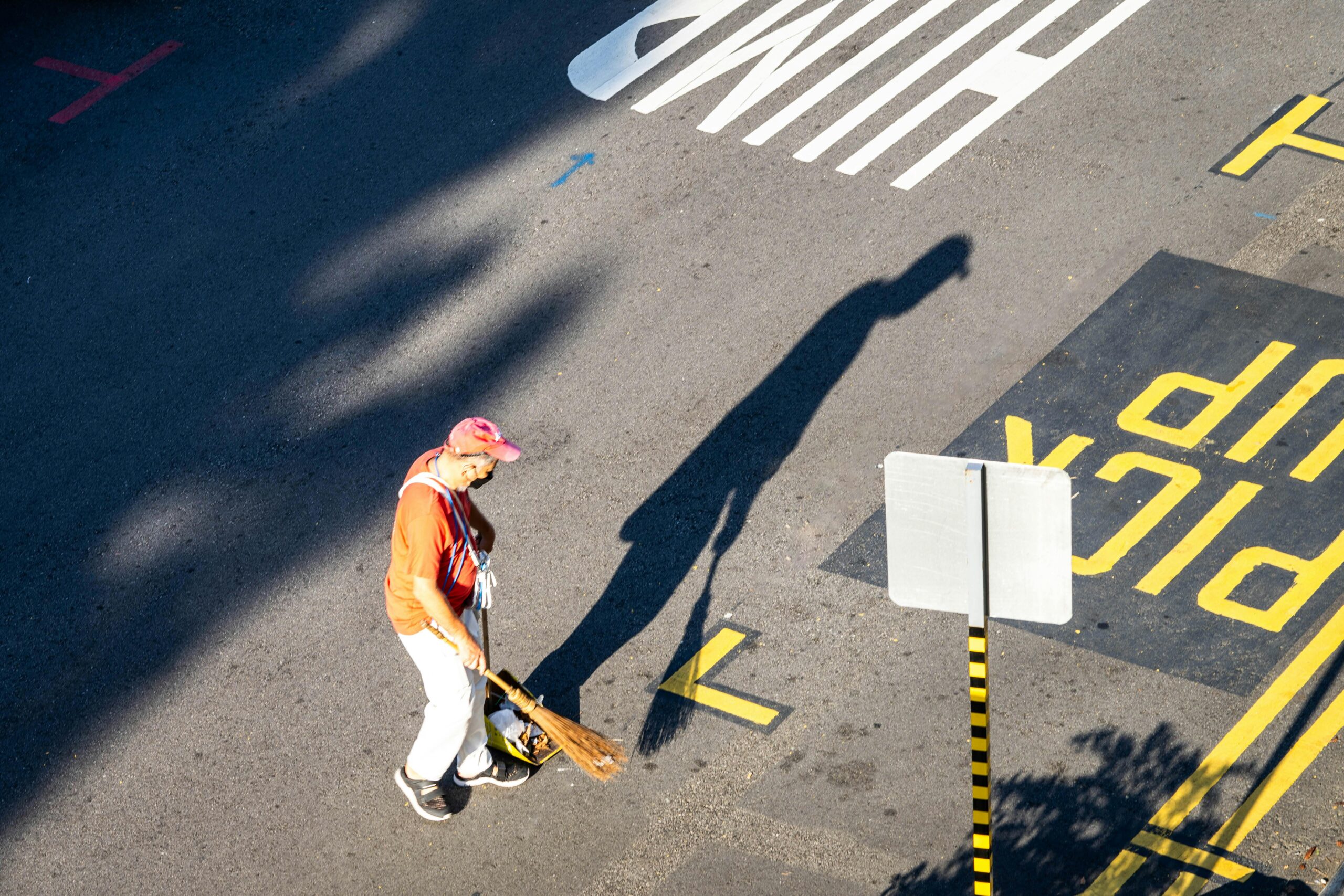 A street cleaner with sweeping tools works under sunlight casting a long shadow on a Singapore city street.